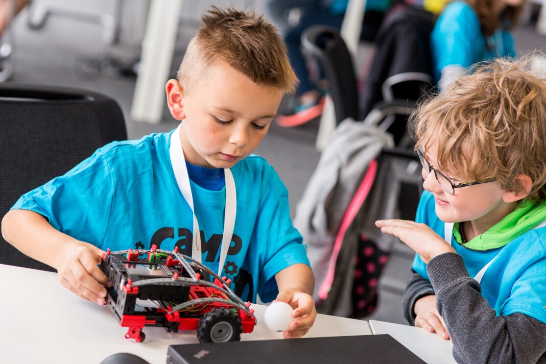 Zwei kleine Jungs blicken auf einen Mini-Roboter, der von einem der Knaben gehalten wird. Vor dem Roboter liegt der Tischtennisball, der transportiert werden soll.