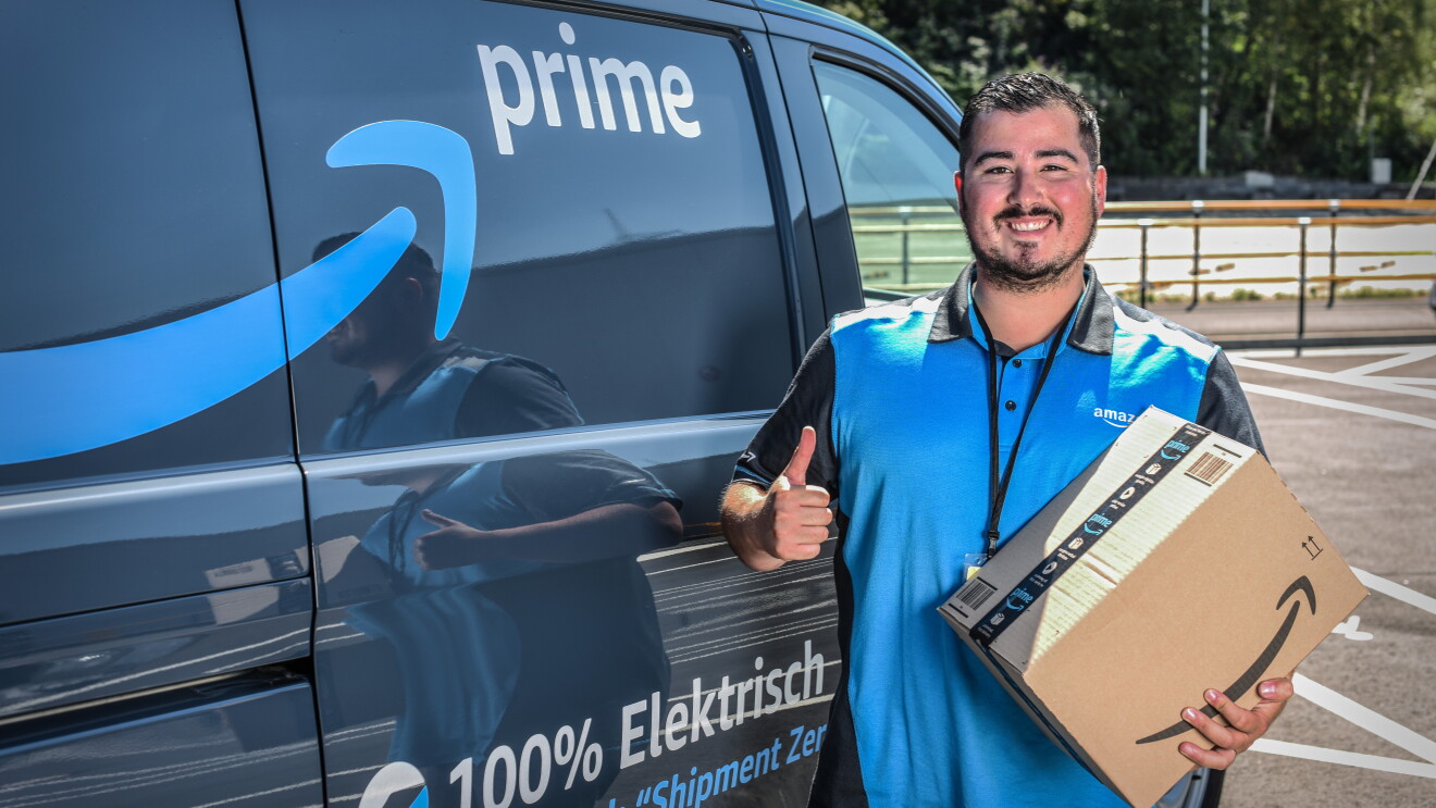 Ein lächelnder junger Mann in Amazon Shirt steht vor einem Prime Lieferfahrzeug. Er hält ein Amazon Paket in der Hand.