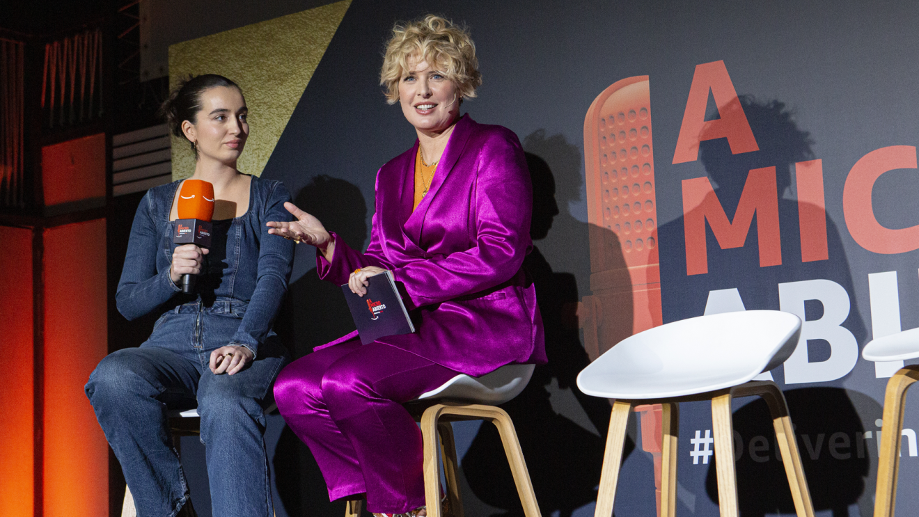 Con el fondo de color azul con el logo de A micro abierto y la etiqueta Delivering Smile, se encuentran dos mujeres sentadas en tabueretes altos. Aparecen la presentadora Tania Llasera (traje chaqueta rosa fucsia, jersey naranja) y otra mujer vestida con mono tejano y con un micro con el logo de Amazon. 