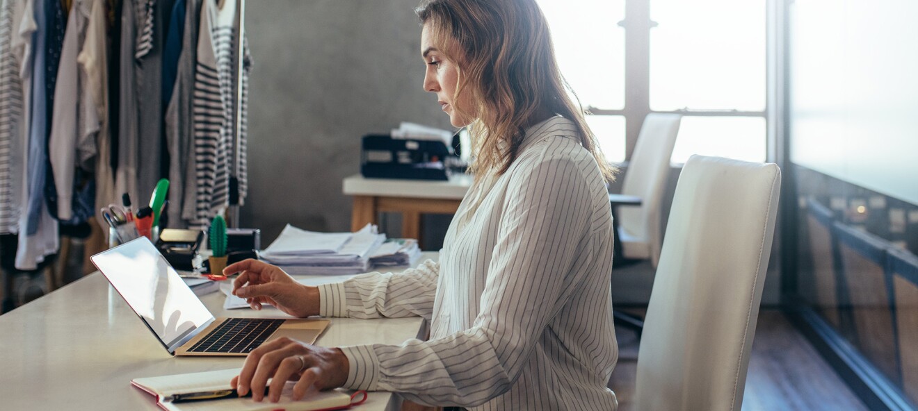 Blonde Frau sitzt am Tisch und arbeitet am Laptop. Im Hintergrund sieht man Stoffe und Kleiderstangen.