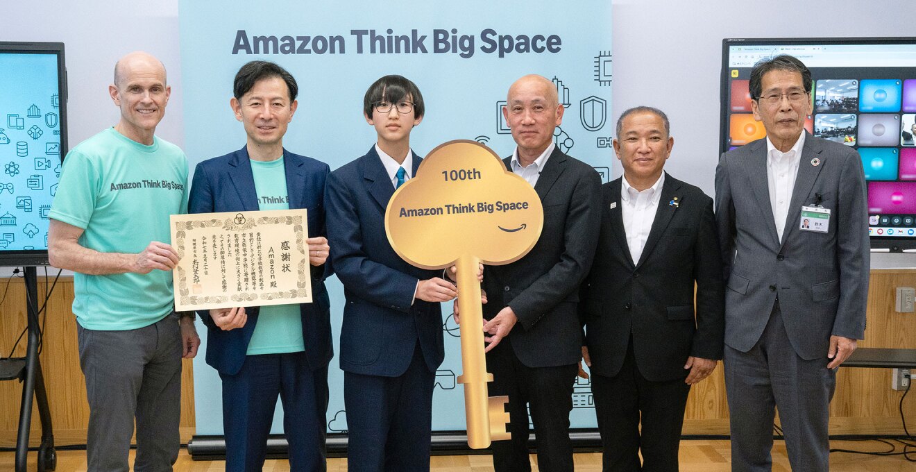 Amazon Think Big Spaceと書かれた背景の前に並ぶ6人の人物。左端の、同じミントグリーンのTシャツを着た2人は感謝状を持ち、中央に立つ制服の生徒とダークグレースーツの人物は金色の大きな鍵型オブジェを持っている。右端の2人は黒とグレーのスーツ姿。鍵型のオブジェには「100th Amazon Think Big Space」と書かれている。