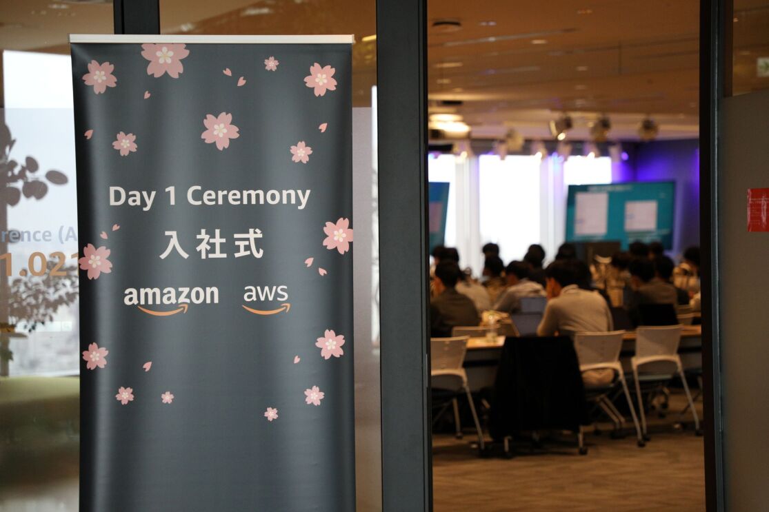 桜模様や「入社式」という文字が書かれている黒いバナー。その後ろの部屋にはたくさんの人が椅子に座っている