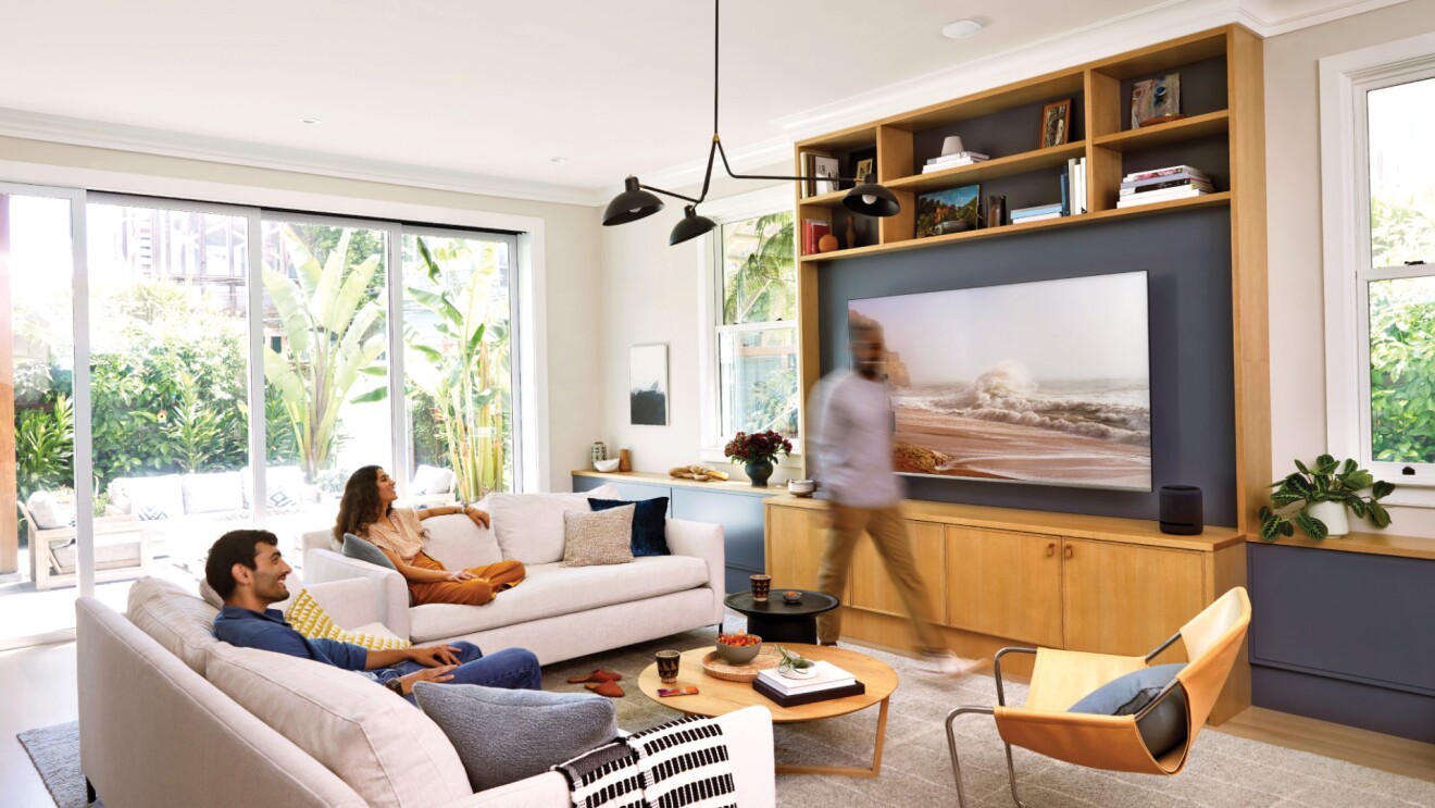 An image of three people interacting in a living room. Two of them are sitting on a couch and the third person is walking by the TV. 