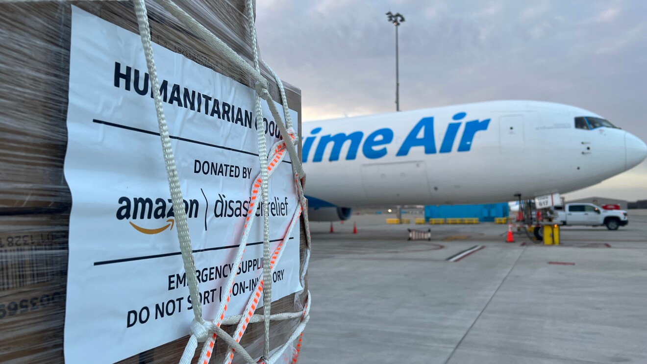 Un palé de ayuda humanitaria en primer plano, de color marrón, con plástico protector, cuerdas y un cartel que pone en inglés Ayuda humanitaria donada por Amazon. De fondo y desenfocado se ve un avión blanco en una pista con las letras en azul Prime Air. 