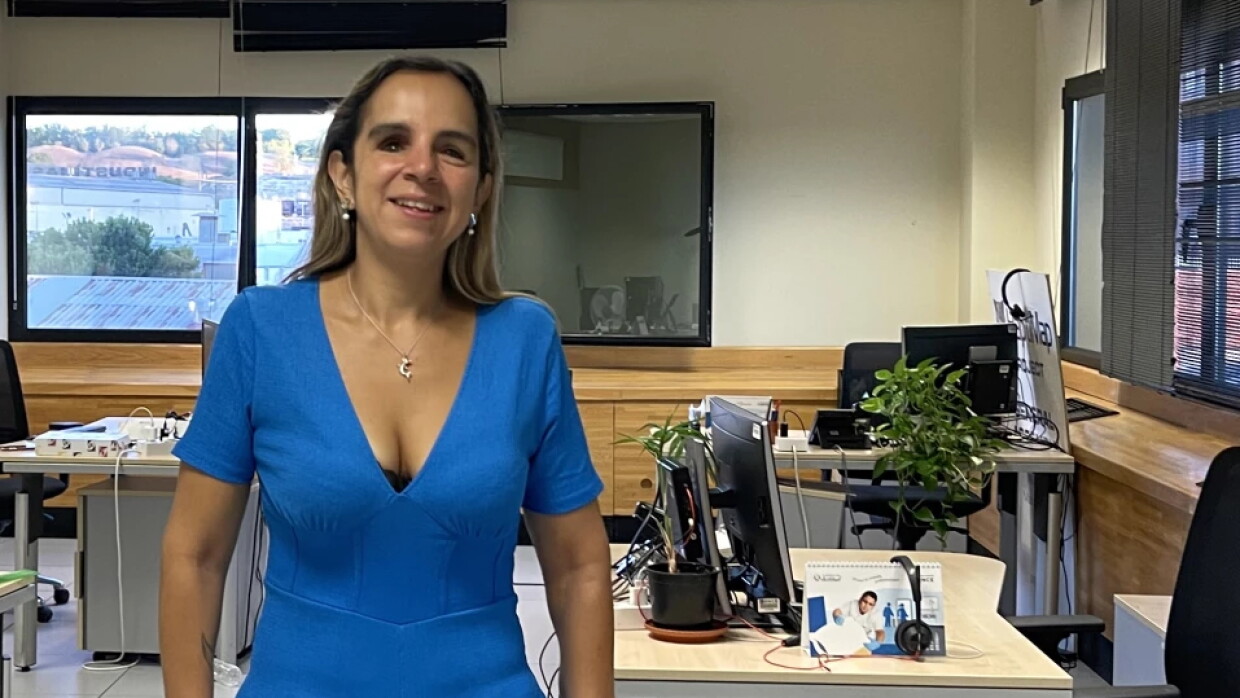 Karina Ramírez Torres se encuentra de pie en su oficina. Lleva un mono de color azul, un reloj inteligente en la mano izquierda, lleva pendientes, un collar y el pelo largo castaño. De fondo se ven cuatro mesas vacías con sus sillas correspondientes y sus ordenadores. Hay dos ventanas por las que entra luz natural.