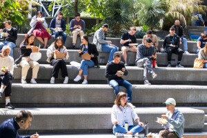 Several Amazon employees sitting on the concrete stairs outside of an Amazon building in Seattle. Many of them are seated in groups, eating lunch from takeout containers.