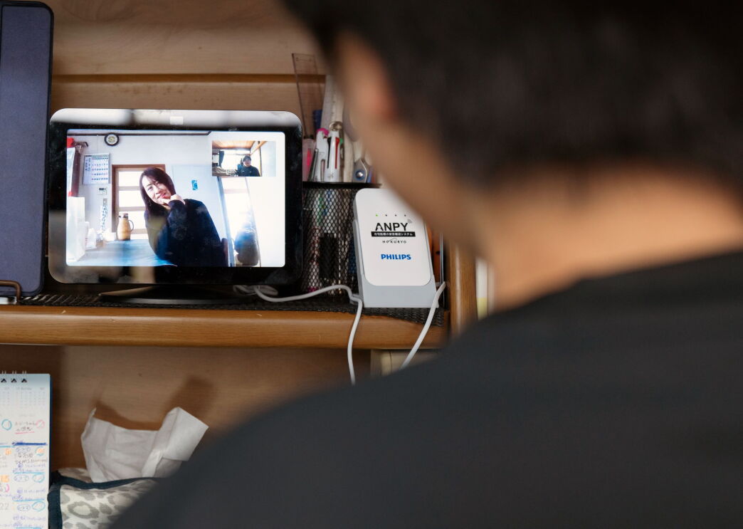デスクのラックに置かれているデバイス（Echo Show 8）の画面に女性が映っている。画面の手前に人物（力斗さん）