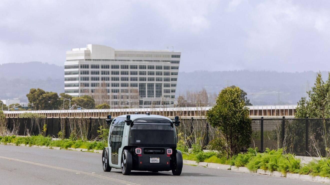 An image showing the Zoox robotaxi opening it's doors on both sides of the vehicle to welcome passengers. There is a passenger entering the vehicle while holding her cellphone and a canned drink. The robotaxi is teal and black and the doors are on a sliding function.