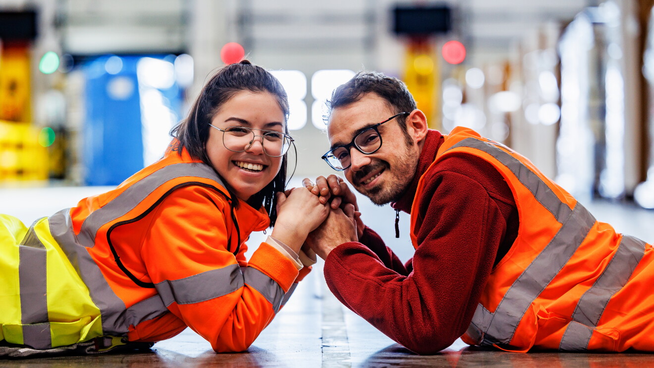 Marta y Roberto posando en MAD6 para la sesión de fotos del post que tenemos entre manos. Los dos están estirados en el suelo. Boca abajo los dos con los codos en el suelo y agarrándose las manos. Miran a cámara. Ella lleva media melena castaña con una coleta alta y el pelo medio suelto. Los dos llevan gafas y él barba y bigote de dos días. Ella una chaqueta de color naranja de seguridad y un chaleco y él un chaleco con un polar de color rojo debajo. De fondo desenfocado un pasillo de un centro logístico. 