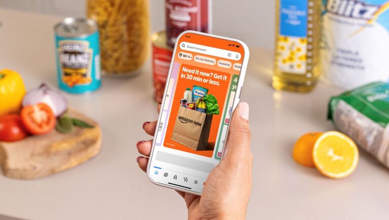 Person holding their phone displaying Amazon Now with groceries on a table in the background.