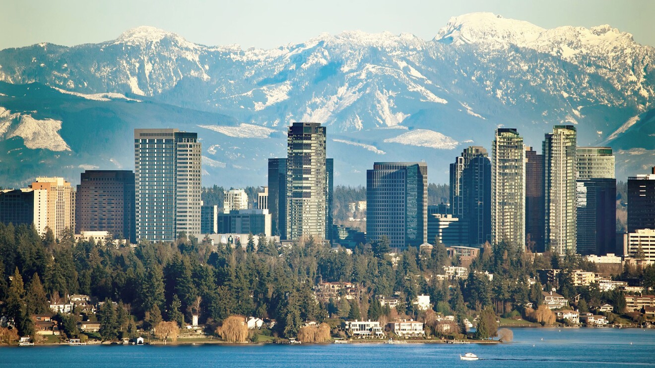 Panorámica del skyline de Bellevue, en Seattle, Washington. Enb primer plano el lago, después muchos árboles y edificios de pocas plantas, en tercer plano muchos rascacielos. De nuevo árboles y de fondo una montañas nevadas.