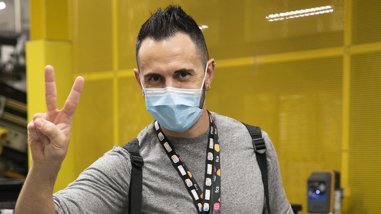 PJammin en BCN1, El Prat de Llobregat. Un asociado mirando a cámara que realiza el gesto de victoria con los dedos de la mano derecha. En la mano izquierda tiene un reloj. El pelo lo lleva de punta y con gomina. Una camiseta de pijama de manga larga pero recogida por el codo y unos pantalones azules con cuadros de color morado. Lleva dos badges y una mochila colgada. 