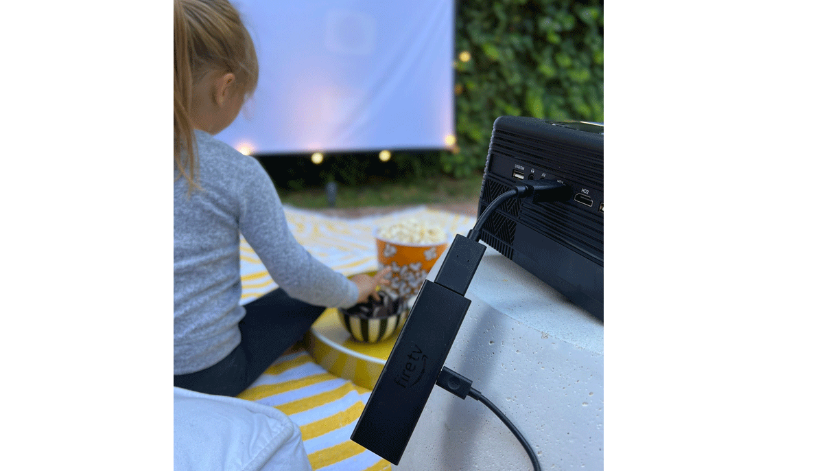 An image of an Amazon Fire TV Stick 4K Max connected to a projector. In the background is a girl eating cookies and popcorn in a backyard.