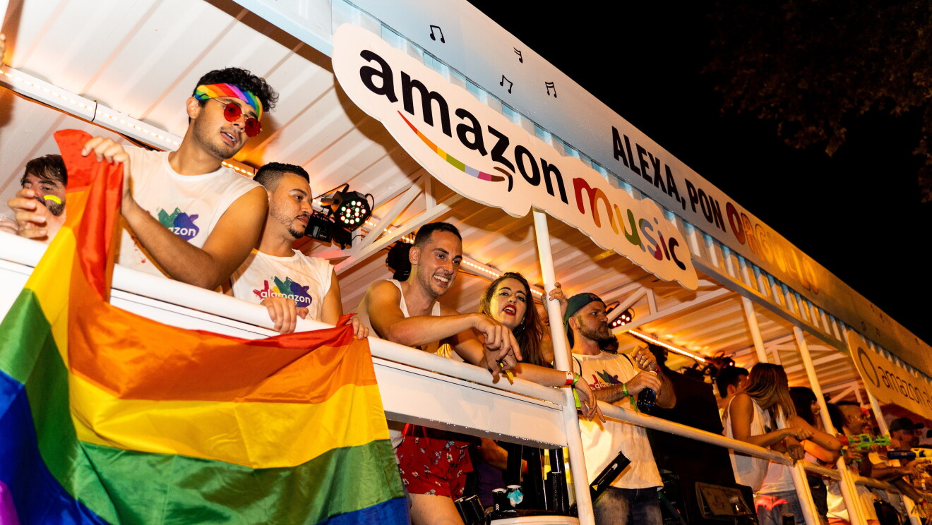 Carroza de glamazon durante el desfile del Día internacional del orgullo LGTBI en Madrid. Ya es de noche y dos chicos con las camisetas de glamazon cuelgan la bandera del arcoiris en la carroza. La carroza está llena de gente que observa a los asistentes al desfile que se encuentran en la calle. En la carroza está el logo de Amazon Music y el lema Alexa, pon orgullo. 