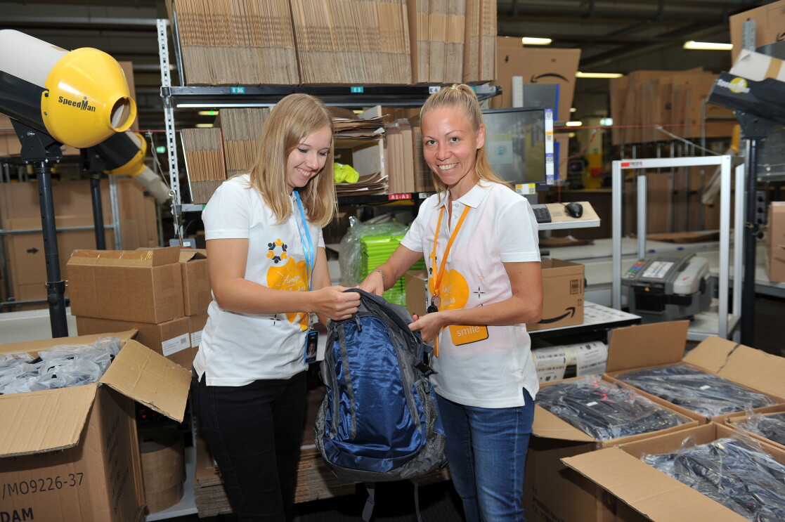 Zwei MItarbeiterinnen von Amazon, beide tragen ein weißes Poloshirt mit orangenen Herzaufdruck und der Beschriftung "Amazon Volunteer" stehen an einer Packstation, umgeben von Kartons. Sie halten in der Hand einen blauen Rucksack, den sie gerade beide mit verschiedenen Untensilien befüllen.