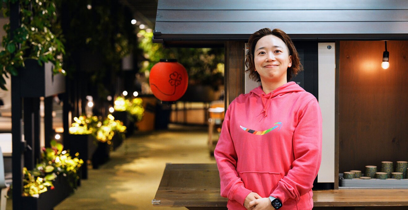 提灯とライトで装飾された木製ブースの前に人物が立っている。レインボーカラーのAmazonロゴが入ったピンクのパーカーを着用している。背景には木製の棚と植物がある