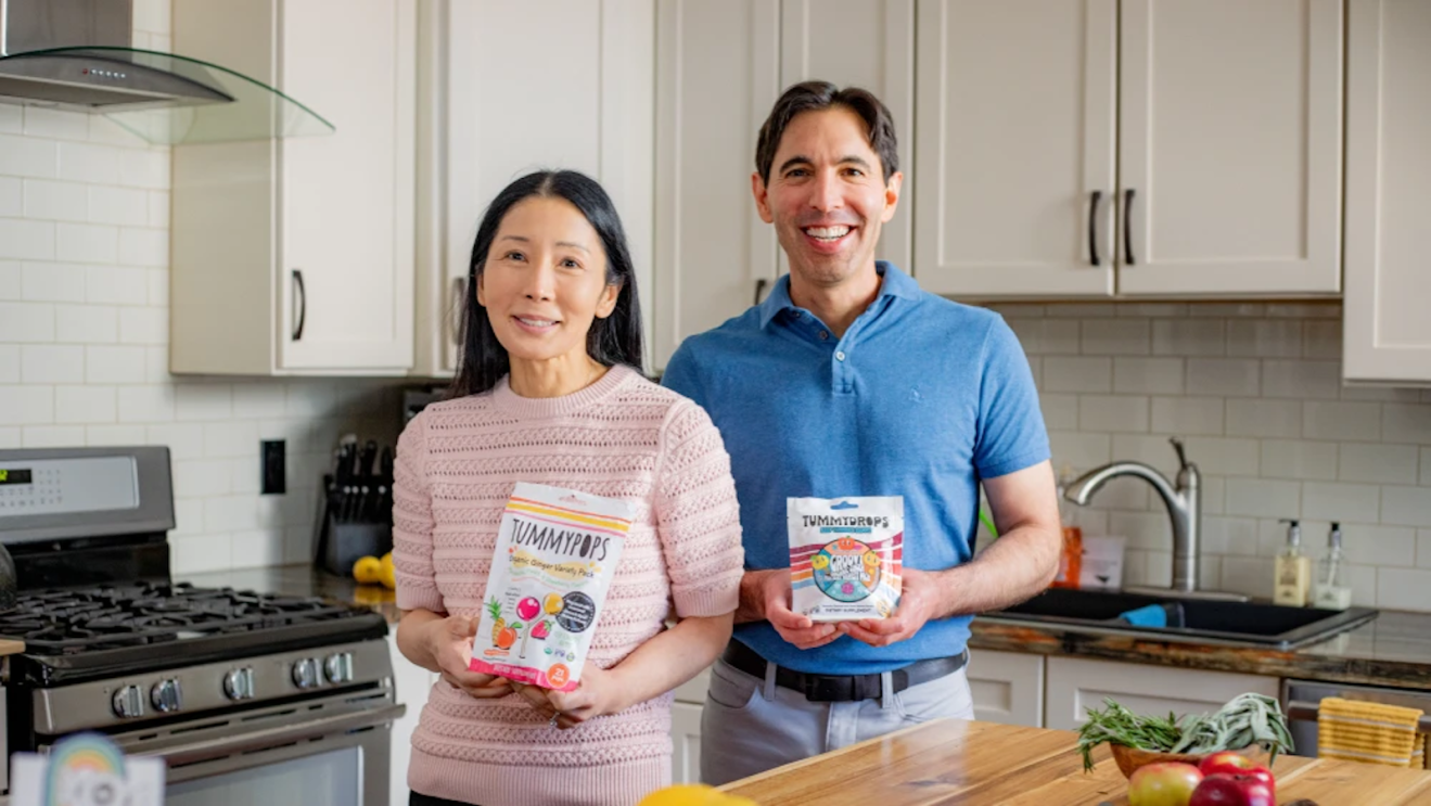Tummy Drops co-founders Dustin and Helen James standing in a kitchen holding bags of their organic lozenge products