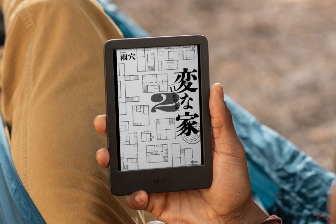 屋外でカジュアルな服装を着た人物が小さな黒いタブレット(Kindle)を持っている。スクリーンにはマンガのタイトルページ「変な家2」が表示されている