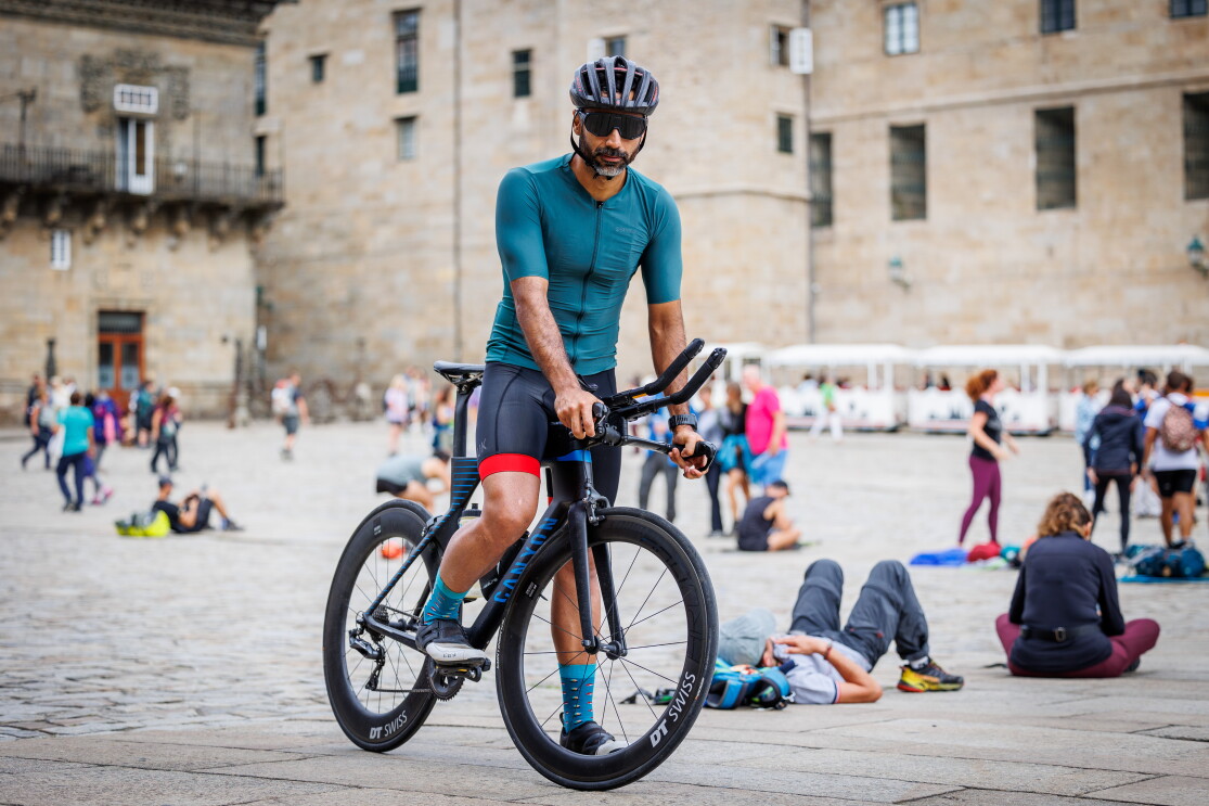 Orlando Aguilera, trabajador del centro logístico de Barcelona. En la plaza del Obradoiro de Santiago de Compostela está Orlando encima de una bicicleta carretera. Apoya las manos en el manillar. Va con el casco puesto, gafas de sol, una camiseta de color azul, pantalones cortos y unas zapatillas de color negro. . Detrás de él está gente sentada en la plaza y yn tren turístico de color blanco. 