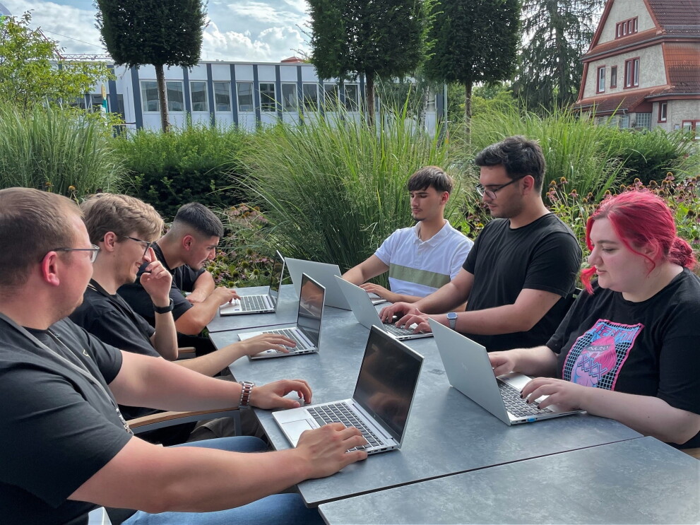 Die ersten Auszubildenden bei AWS sitzen lächelnd draußen an einem Tisch mit Laptops.