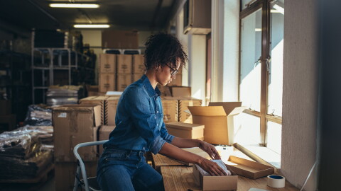 A woman sits on a step stool as she packages a product in a kraft box. 