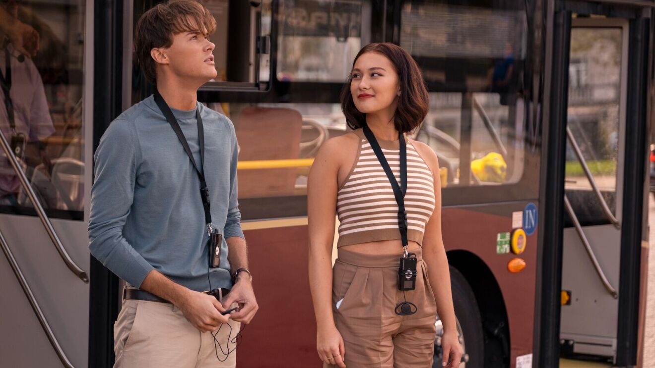 A scene from The Summer I Turned Pretty with two people with lanyards standing in front of a bus, dressed casually for summer