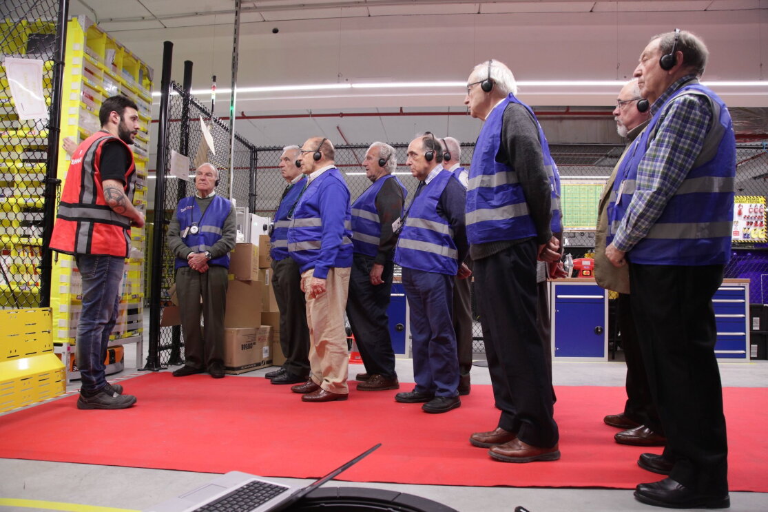 Nueve ingenieros jubilados durante la visita a un centro logístico. Todos ellos visten con un chaleco y escuchan atentamente las explicaciones del ingeniero que lleva un chaleco rojo. 