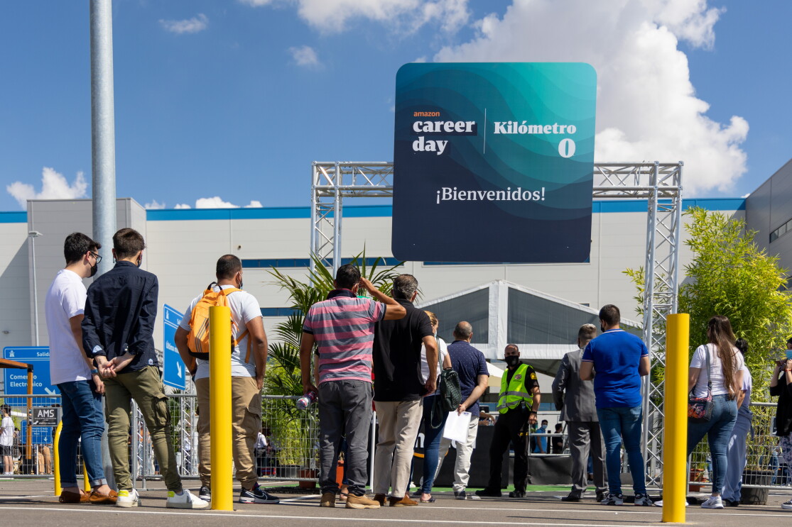 30 000 personas solicitaron un puesto de trabajo en Amazon durante el Career Day. En primer plano un grupo de 15 personas de espaldas a la cámara haciendo cola para participar en el Career Day de Amazon 2021. De fondo el centro logístico de Amazon en Illescas. La gente está haciendo cola, en la entrada, delante de un cartel que está apoyado en la parte superior de una columna, En el cartel pone ¡Bienvenido!, kilómetro 0 y Amazon Career Day. Se ve el cielo azul con nubes blancas. 