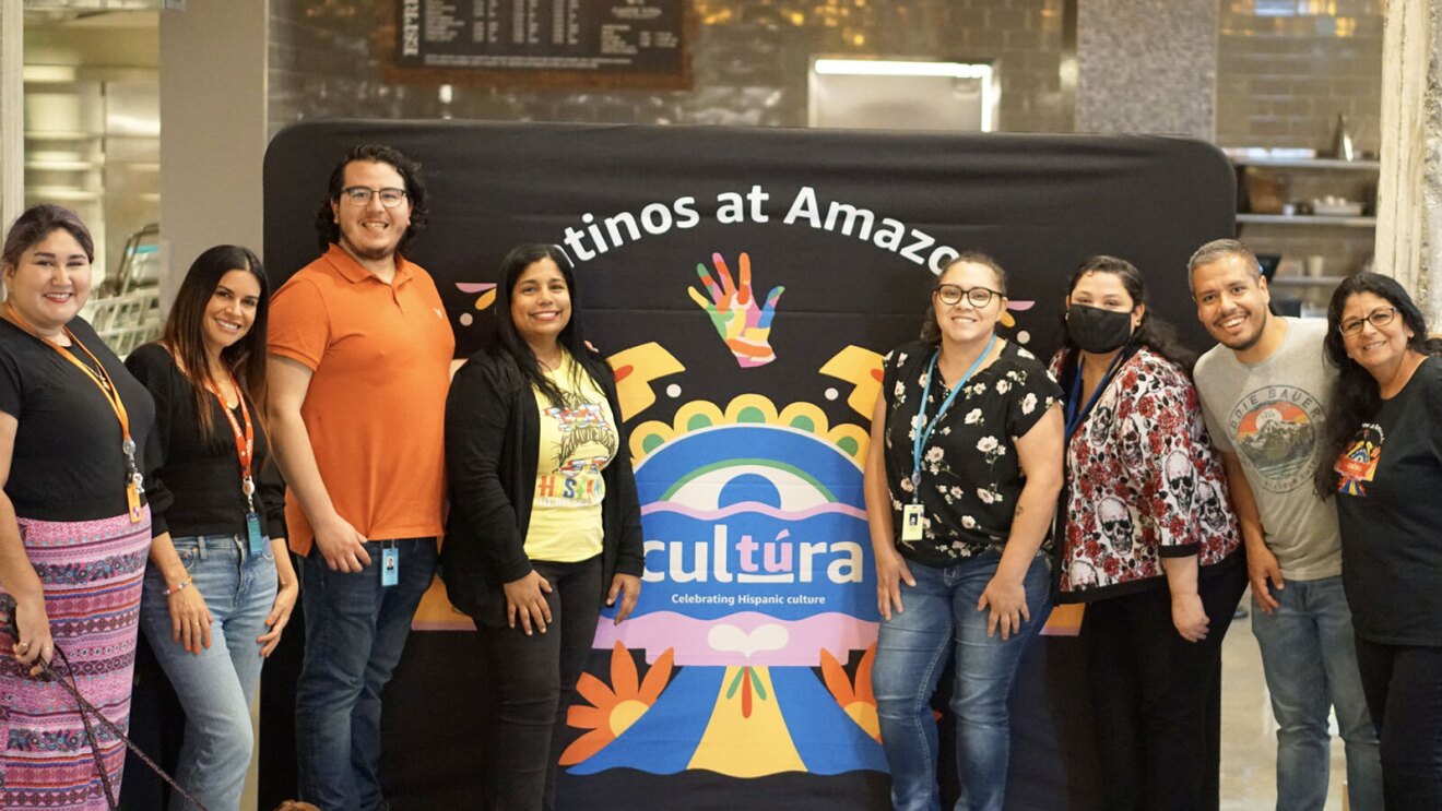 A photo of eight employees standing in front of a Latinos at Amazon cultúra banner. 