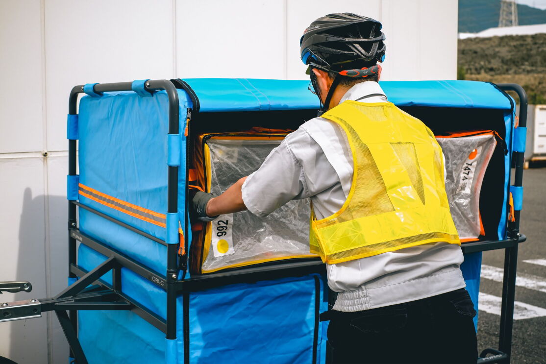 黄色い安全ベストを着てヘルメットをかぶっている男性が大きな荷物をリヤカーの中に積み込んでいる。
