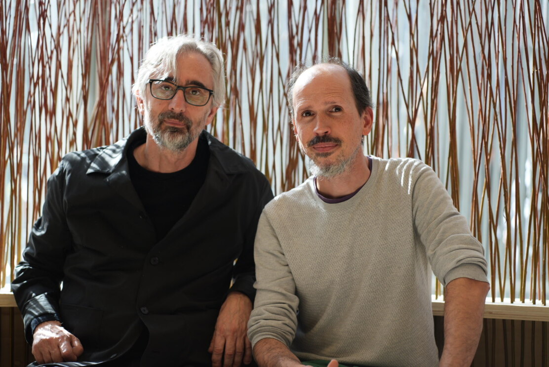 Martín Azúa e Iván Bravo posan en el Madrid Design Festival. Martín tiene pelo canoso, barba, gafas de pasta y lleva puesto un jersey de manga larga negro. Iván tiene barba negra y bigote, y lleva puesto un jersey beige. Ambos están sentados uno junto al otro y miran a cámara sonriendo. 