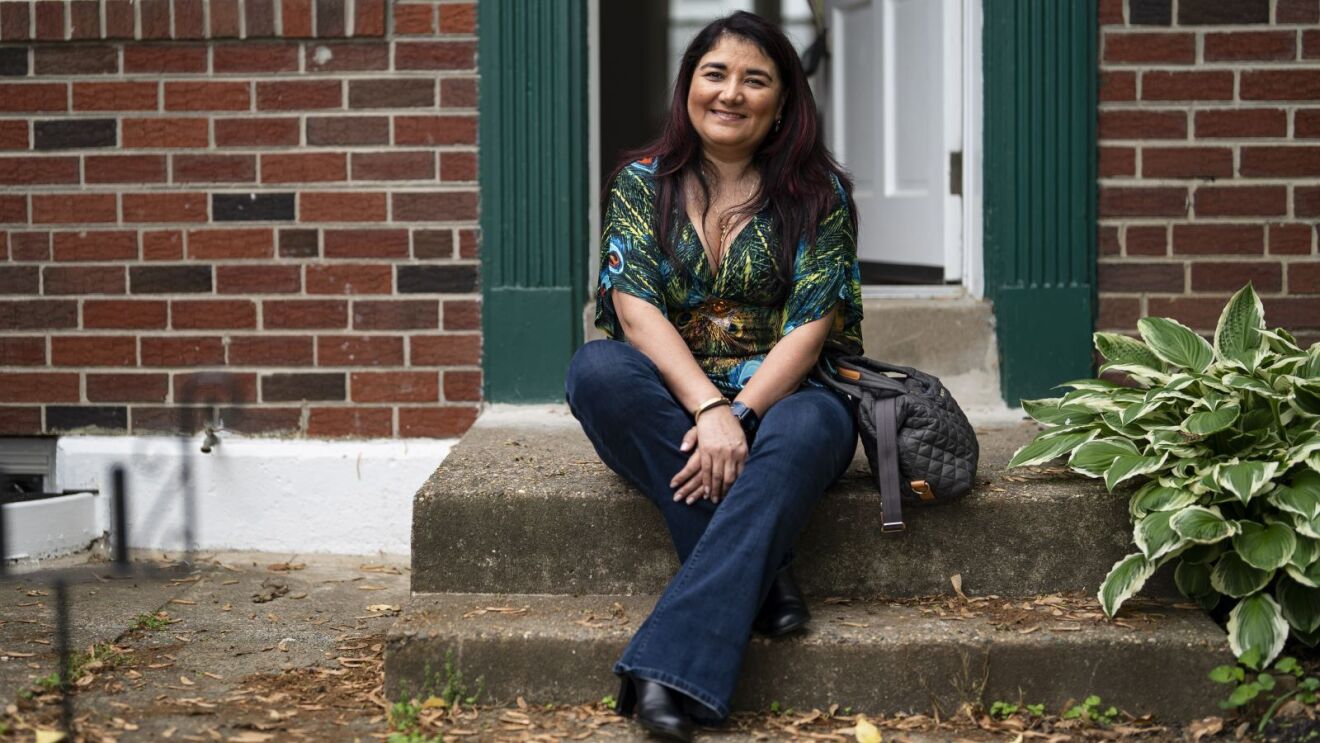 Smiling woman in patterned shirt and jeans sitting outside traditional brick house