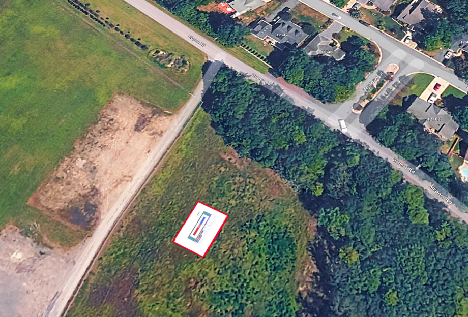Aerial/satellite view of a proposed site outlined by a red rectangle in a cleared grassy area beside a wooded lot. A small building footprint diagram is overlaid inside the red boundary. A road runs along the top with a residential neighborhood of houses to the right and an adjacent graded/excavated area to the left.
