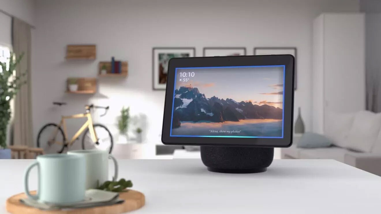 An image of an Echo Show device sitting on a kitchen counter. There are coffee mugs in the foreground on the counter and in the back is a living room space with a bike, shelves, and framed images on the wall.