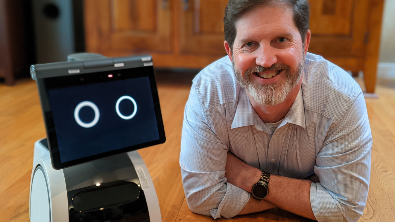 A man lays on the floor and smiles as the camera, next to an Amazon Astro house robot, in a kitchen.