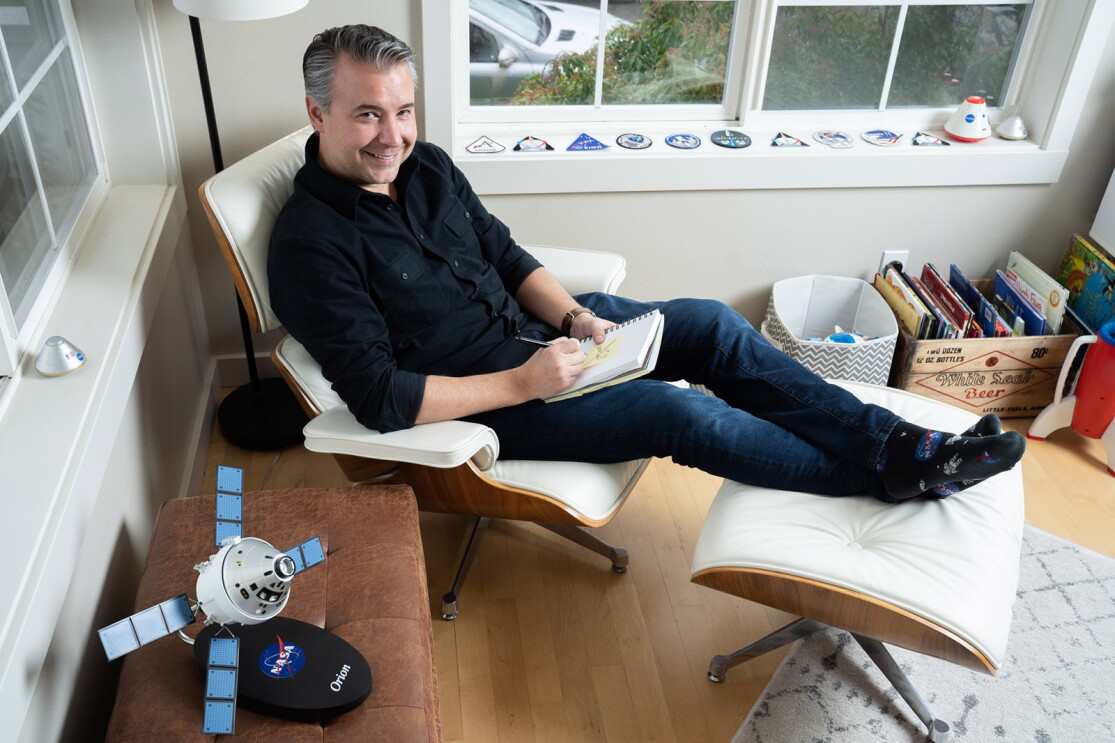 An image of a man sitting in a chair in his office. He is looking up at the camera and smiling while drawing on a notepad and there is a small satellite device on a table next to him.