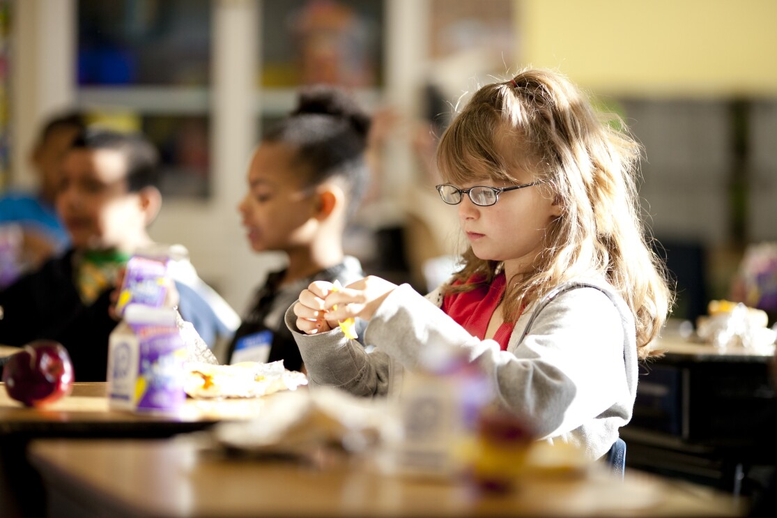 A young girl is opening preparing to eat. Behind her, other students are also eating. Apples and single-serve milk containers are on the desks in front of the students.