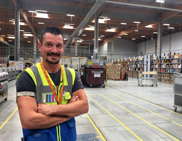 The image of Lionel Boulbes at the facilities center in his yellow vest with boxes and trays in the background. He is a French Amazon associate working at Montmelier facilities center in France. 