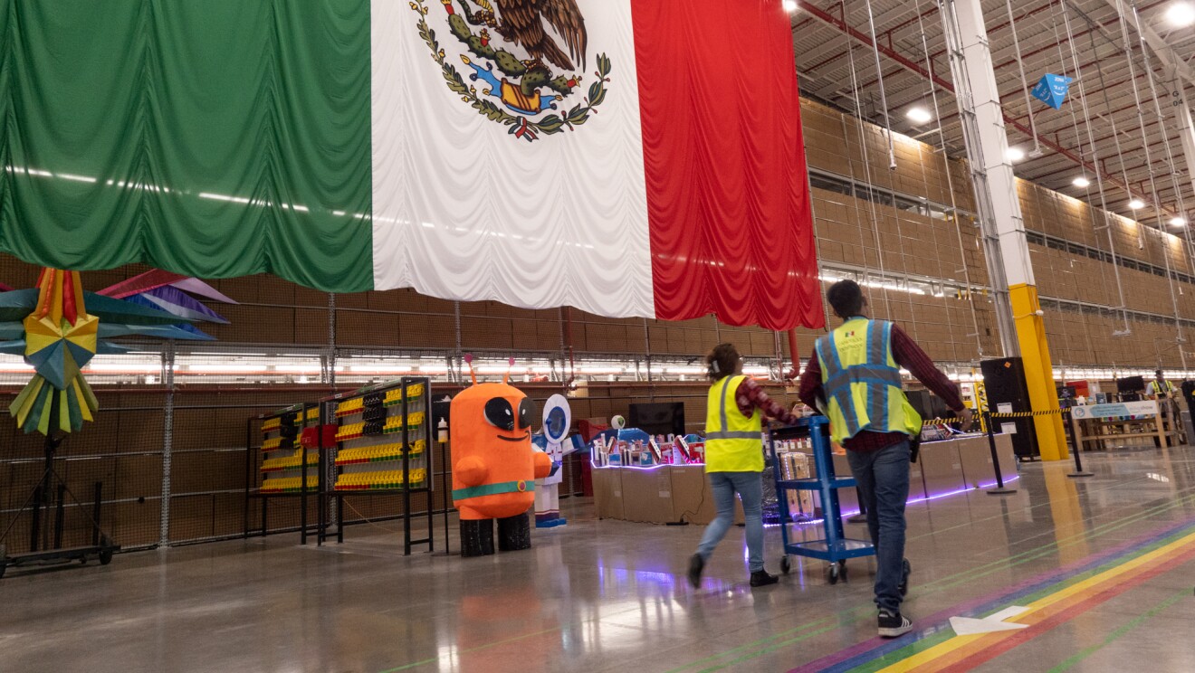 La entrada de un centro logístico de Amazon con una bandera de México  de unos 3x6 metros. Aparece la mascota Peccy que es la encargada de temas de seguridad y dos personas de espaldas con el chaleco amarillo y tirando de una estantería con rueda de unos 90 cm.