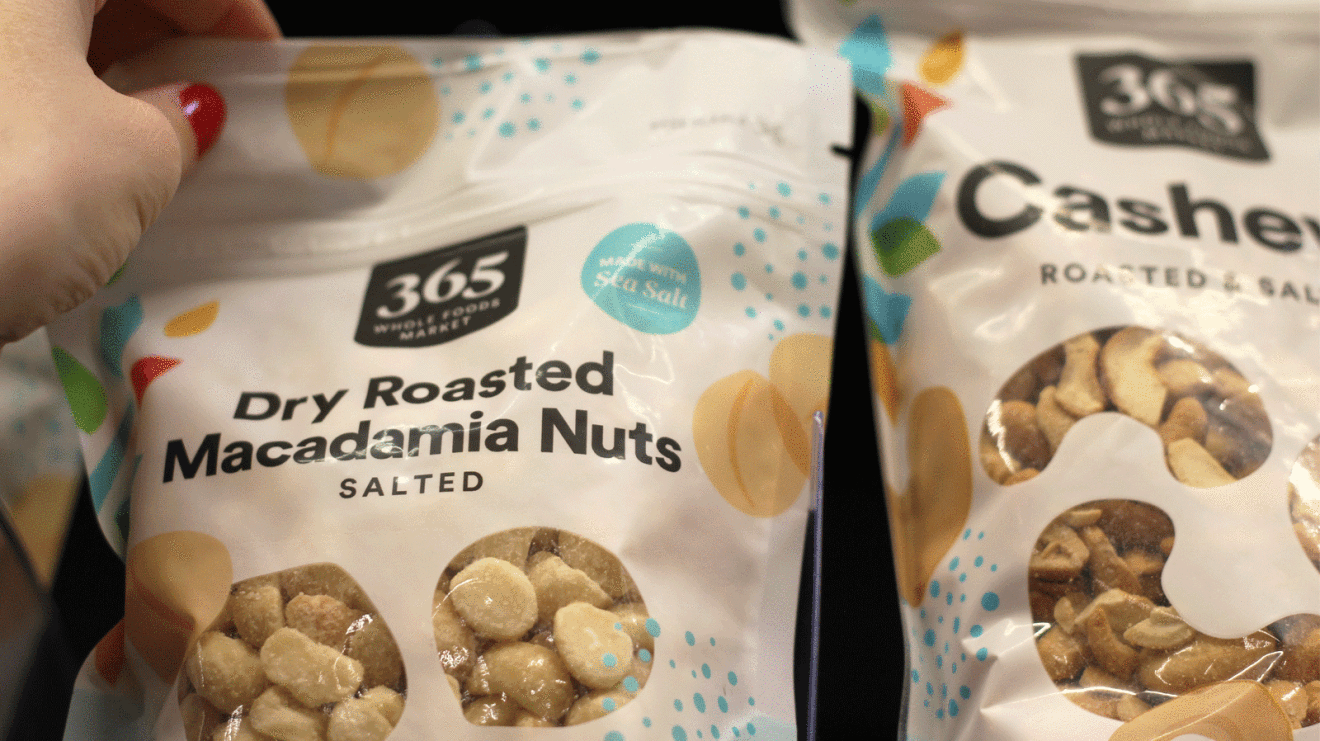 An image of a hand pulling a bag of Macadamia nuts from the 365 Brand off the shelf.