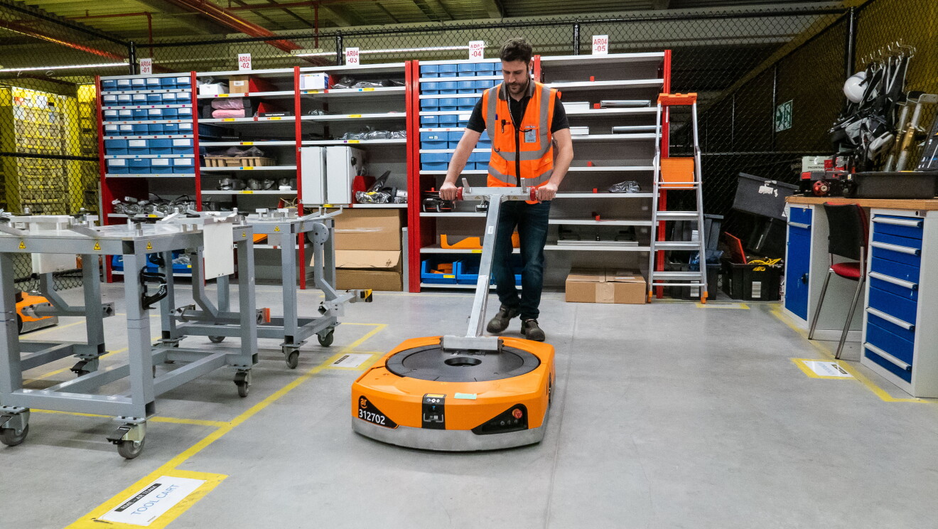 Raúl Cruz, técnico senior de mantenimiento en robótica, manipulando un robot (drive). Vestido con el chaleco naranja, Raúl aparece trasladando un robot con una herramienta específica que sujeta el drive por su parte inferior. De fondo de foto aparecen estanterías propias del lugar de reparación de los robots. 