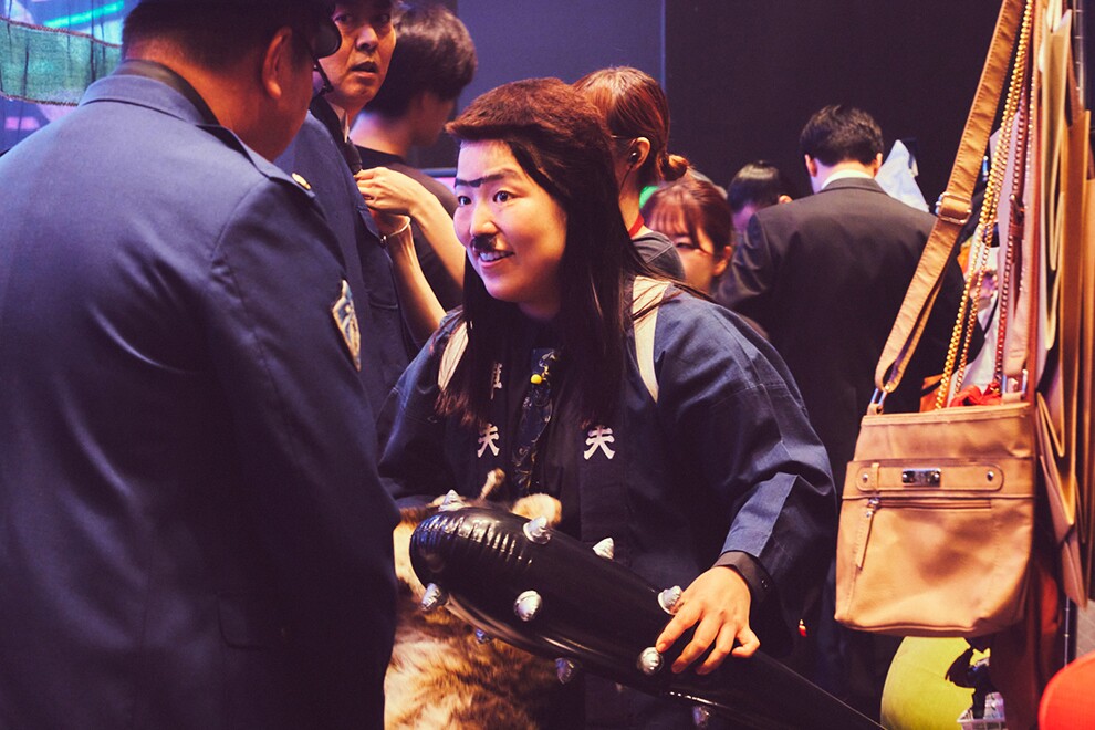 黒い壁の室内に立つ複数の人々。中央にバルーンの金棒を持って紺色の半纏、付け髭、かつらを着用した人物。左側に警官の制服を着た男性の後ろ姿。右側には鞄などの小道具。