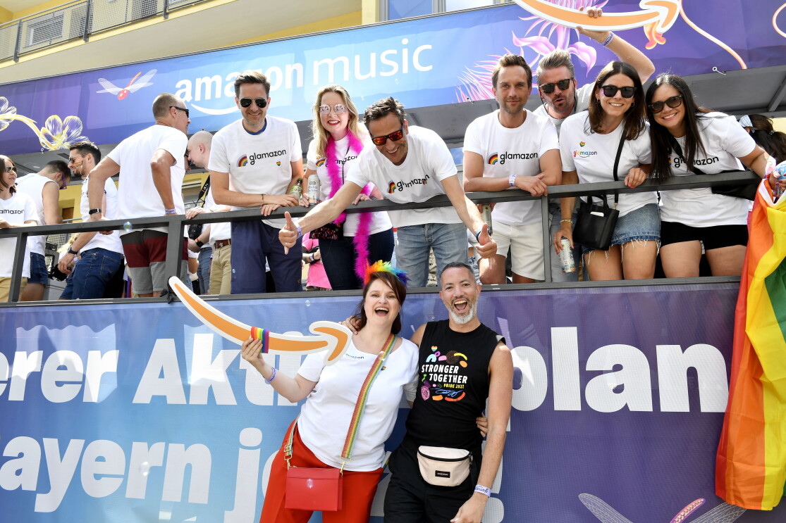 CSD-Truck von Amazon in München: In der oberen Reihe sieht man Rocco Bräuniger mit einer Reihe von Mitarbeiter:innen: Alle Tragen weiße T-Shirts mit der Aufschrift "glamazon". In der Reihe unten stehen eine Amazon Kollegin und ein Kollege Arm in Arm und mit dem Amazon Logo in der Hand.
