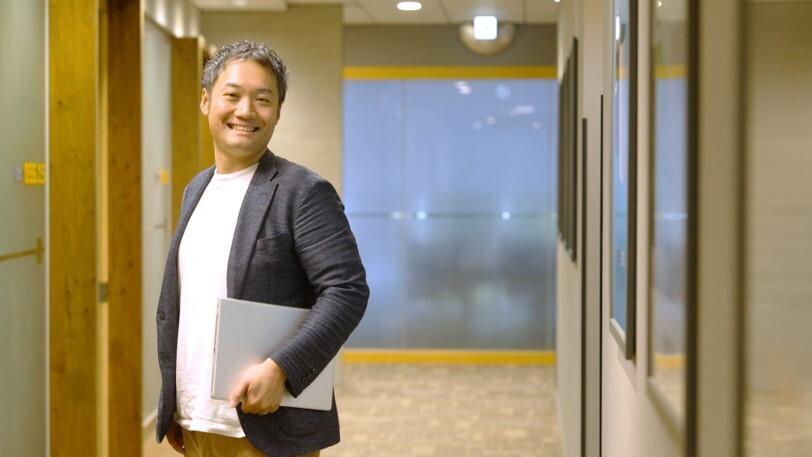 オフィスの廊下に立つジャケットを着た男性、片手にPCを抱えている