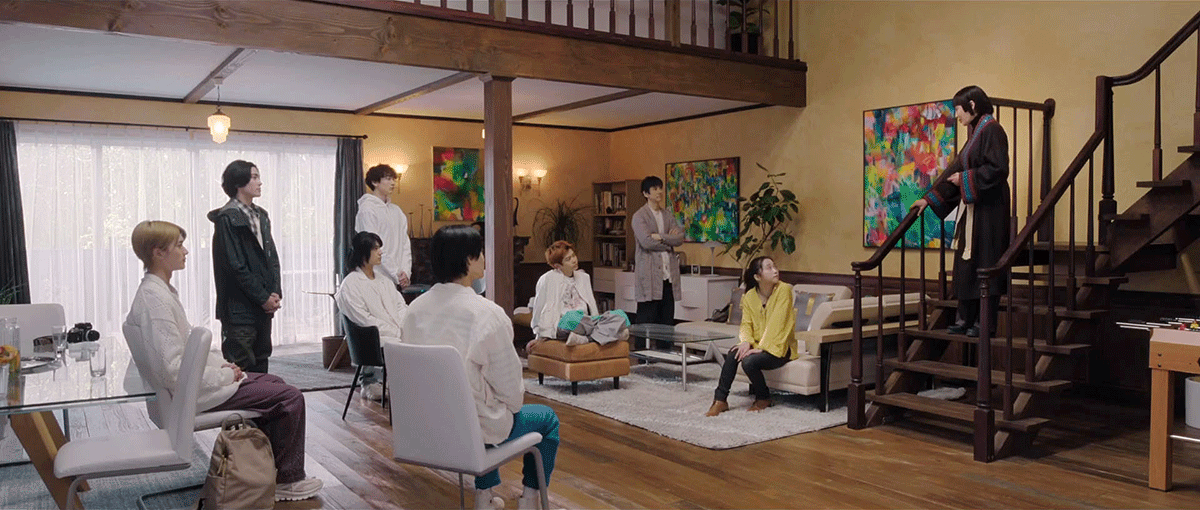 広い室内に集まった9人の人々。右端の階段の途中に立つ、ガウン着用の人物に他全員の視線が集まる。うち5人は椅子やソファに腰掛け、3人は立っている。ベージュの壁にはカラフルな絵画が3点。