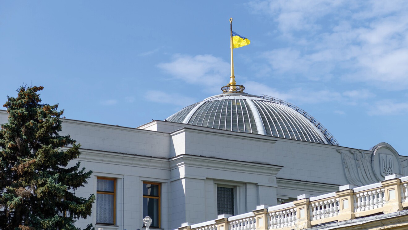 An image of the parliament building in Ukraine. There is the Ukraine flag flying atop the building and a blue sky in the background.