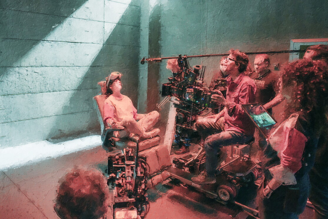 Fotomontaje. Es una fotografía que lleva un filtro que simula un cuadro pintado. Varios empleados trabajando en el rodaje de Reina Roja. Un fondo liso de color azulado con una personas sentada en una silla de hospital y con unas gafas 3D. Delante de ellas un travelling con una persona sentada delante de una cámara y al lado izquierdo la cabeza de un hombre con una cámara. En un segundo plano cuatro personas mirando la imagen. Y sale una pértiga de sonido que no se ve quien le agarra.