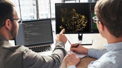 两人坐在办公桌前工作的照片。有一个笔记本电脑设备和桌面设备放在桌子上。