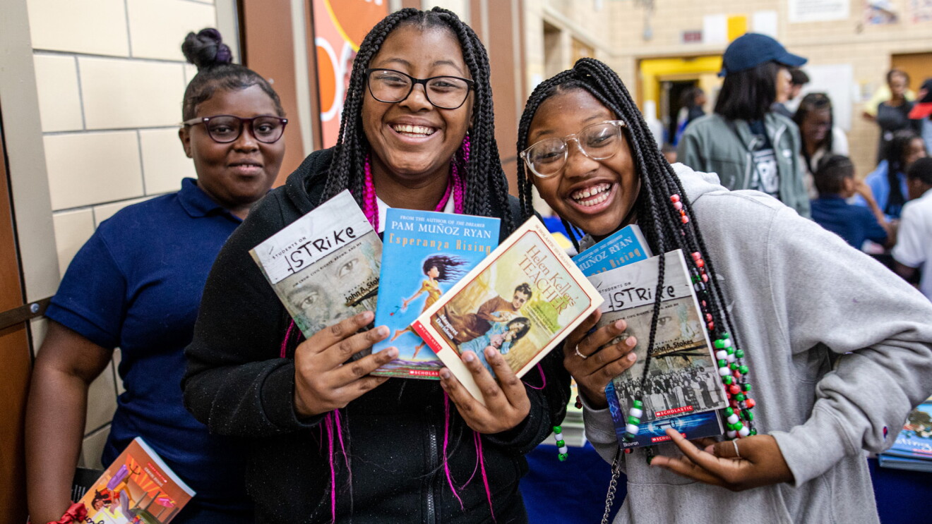 An image of three students smiling at the camera and holding up books. 