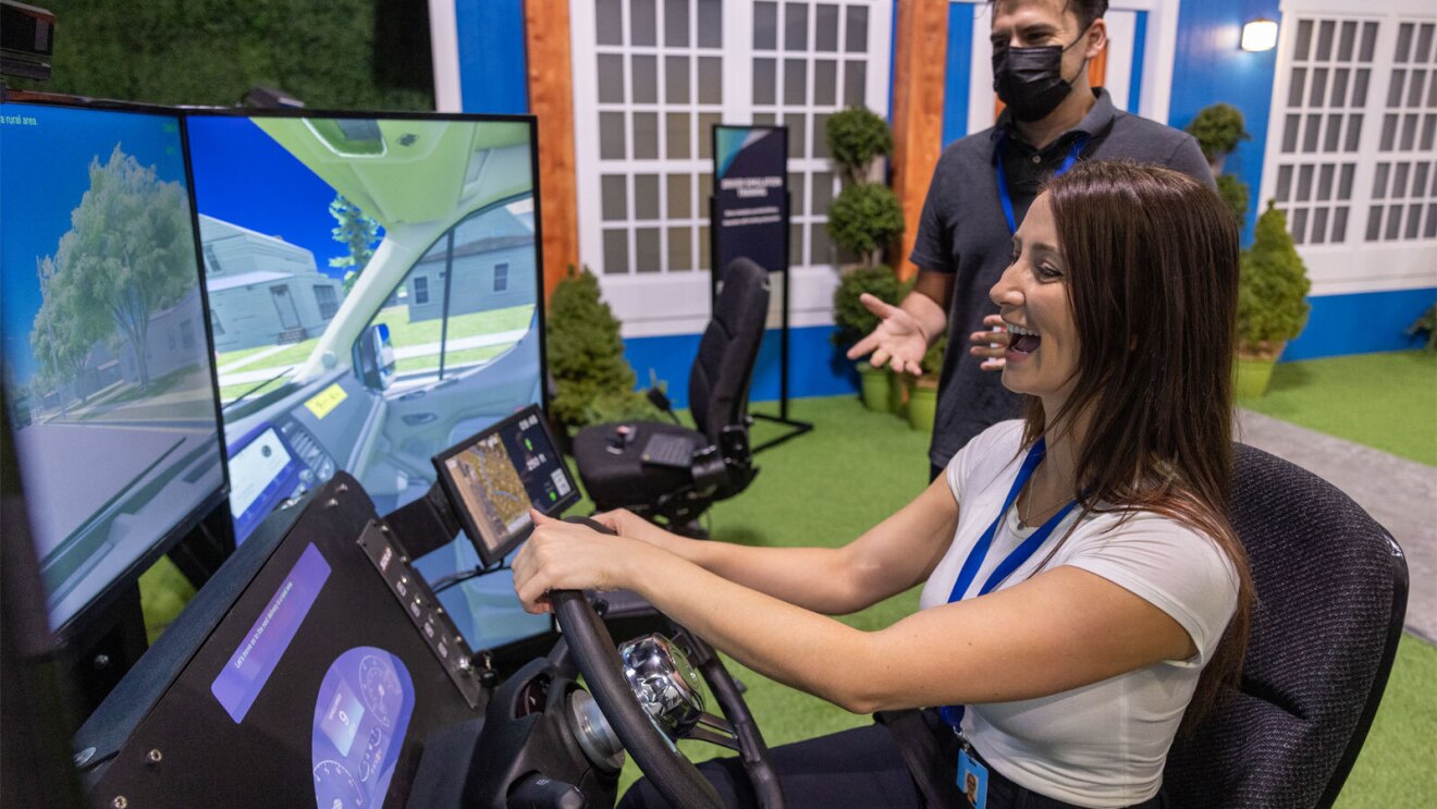 An image of a woman smiling while driving a simulator of a rivian van. She is sitting in a mock driver's seat, holding the steering wheel and looking at three large screens in front of her that show a simulated neighborhood road.